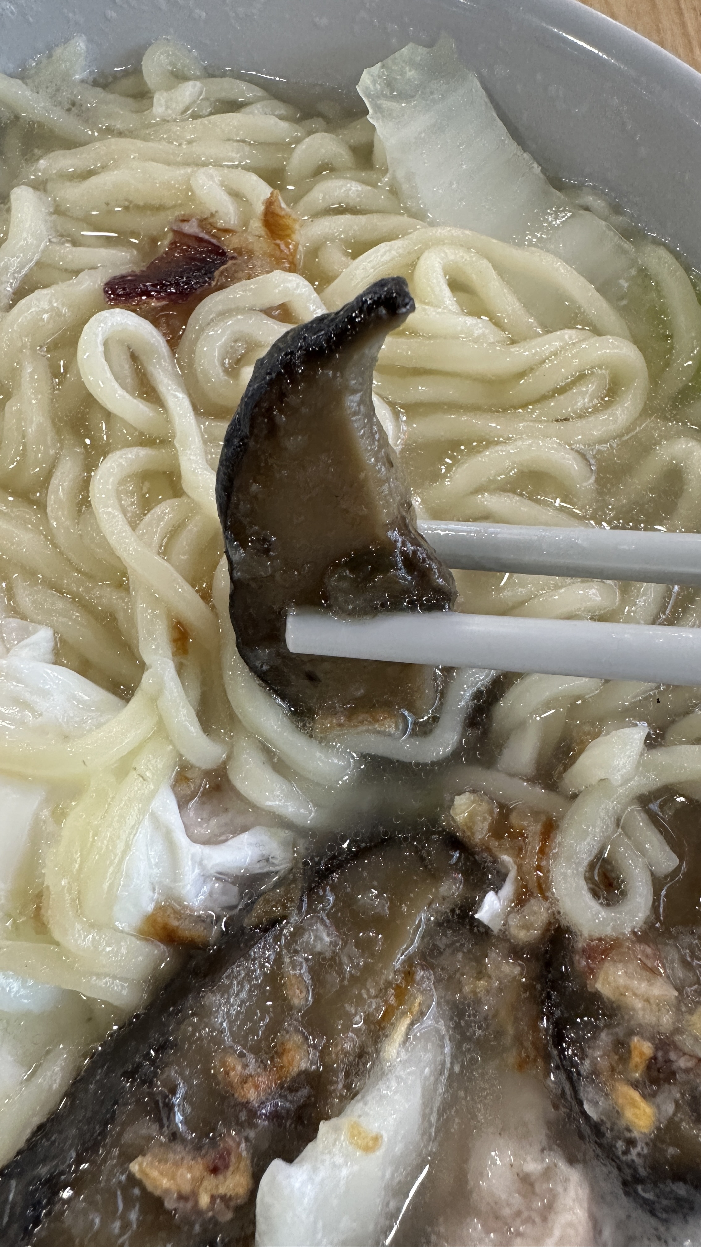 A picture showing a pair of chopsticks holding up to a piece of stewed shiitake mushroom on a bowl of youmian soup.